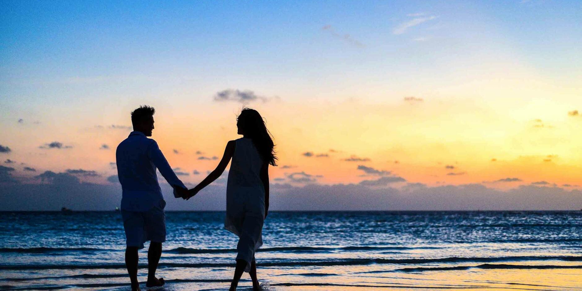 man and woman holding hands walking on seashore during sunrise