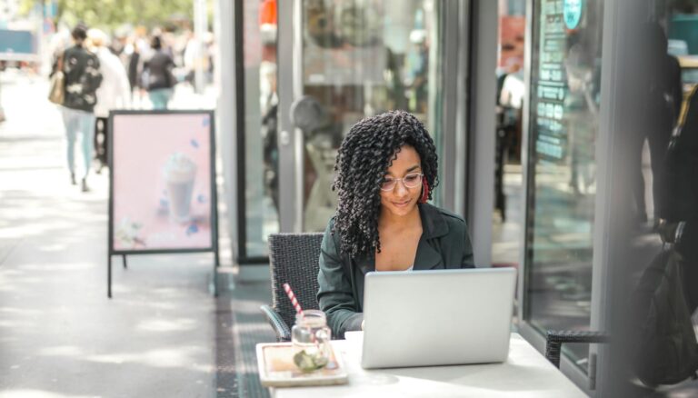 ethnic young woman using laptop while having tasty beverage in modern street cafe
