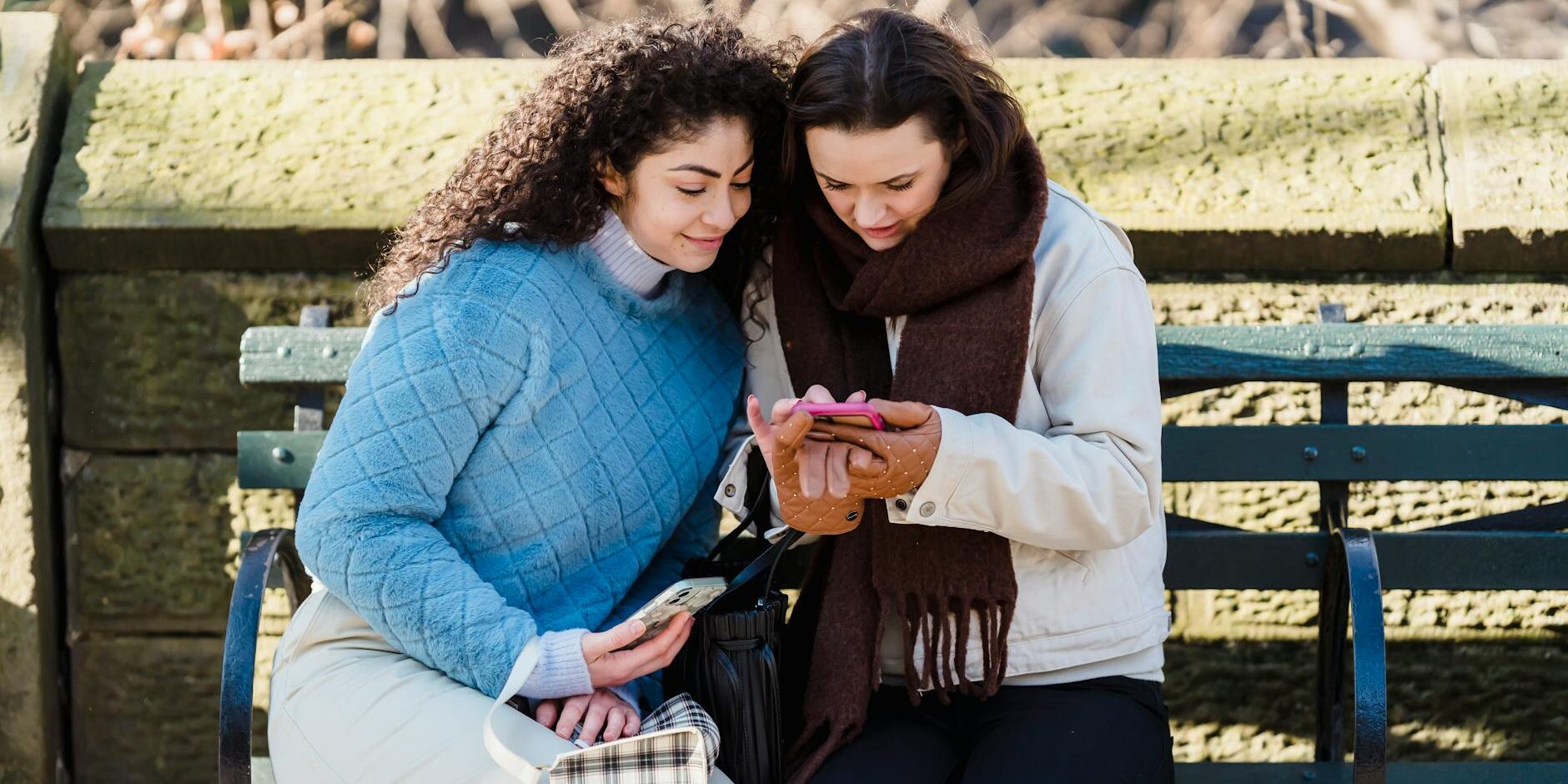 trendy women using smartphone together on bench in park