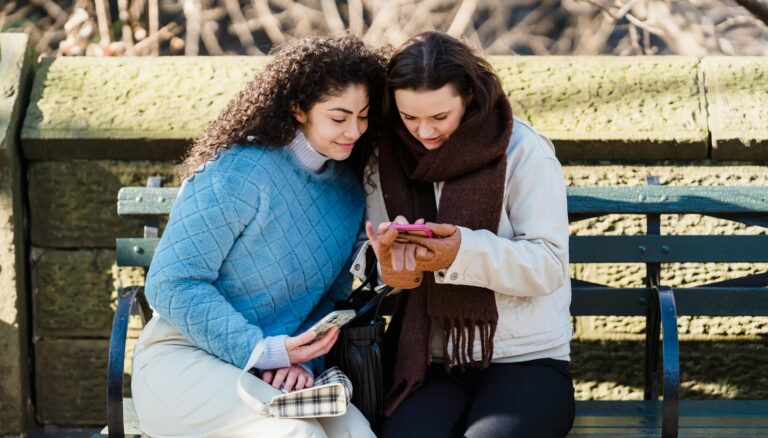 trendy women using smartphone together on bench in park