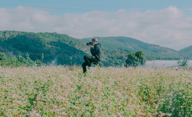 man sitting in the middle of flower field using black dslr camera